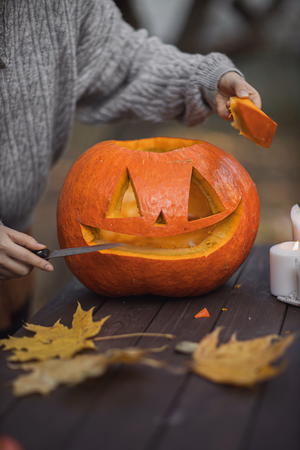 A person in a gray sweater cutting a triangle eye out of a pumpkin with a knife on an outdoor wooden table scattered with yellow leaves