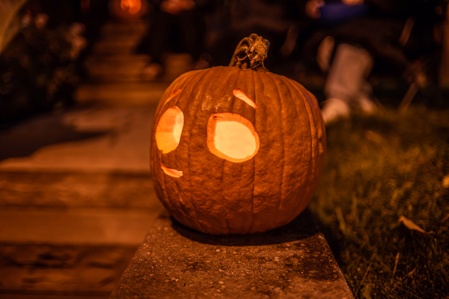 A lit jack-o'-lantern sitting on the edge of front steps at night, a trick-or-treater's costume blurred in the background