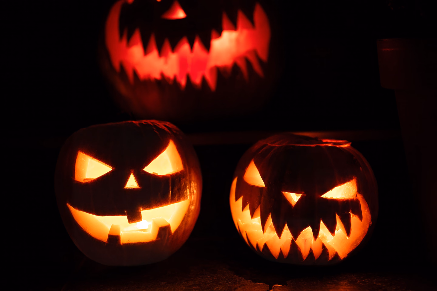Three carved jack-o'-lanterns with angular eyes and toothy grins glowing orange and red in total darkness