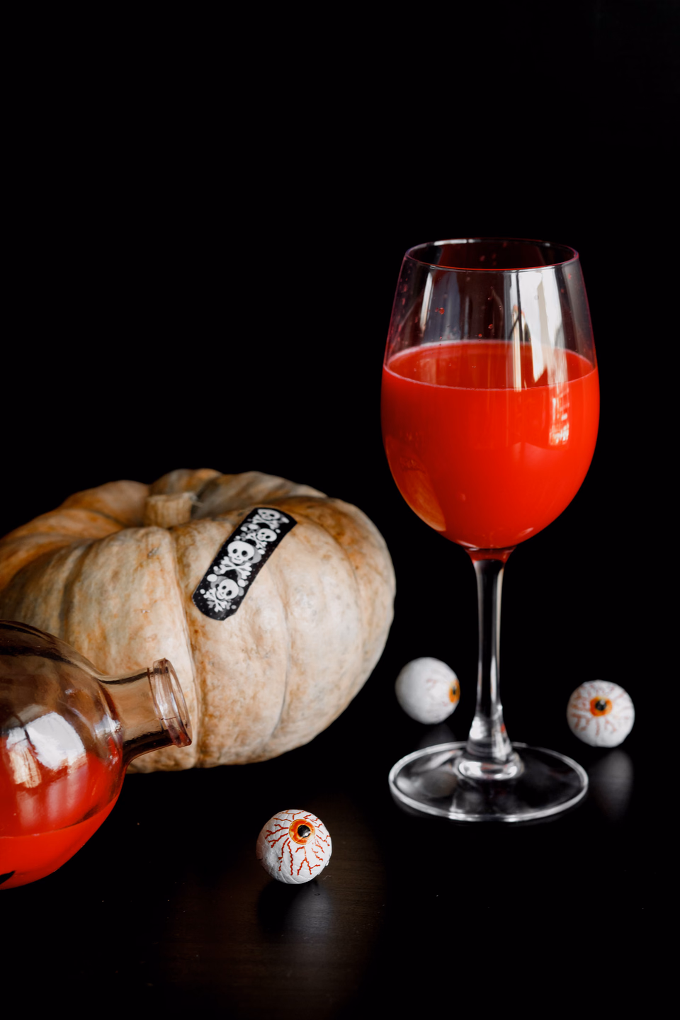 A stemmed glass of bright red liquid on a black surface beside a pale pumpkin, a small bottle of red liquid, and three candy eyeballs