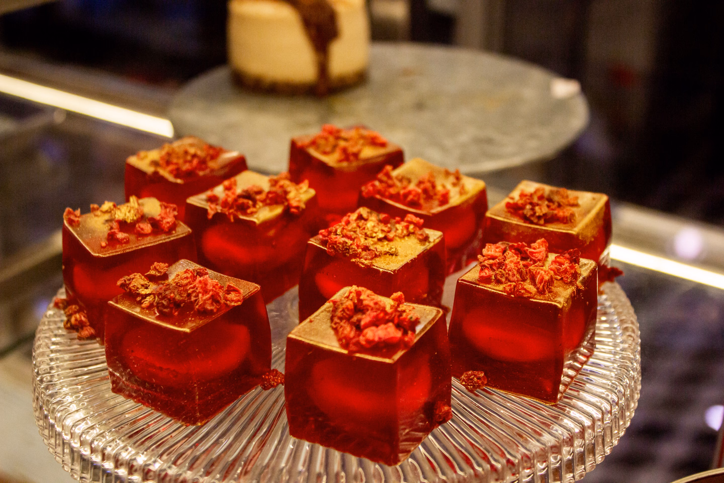 Neat cubes of clear red gelatin topped with crushed dried berries and gold flakes, arranged on a cut-glass platter