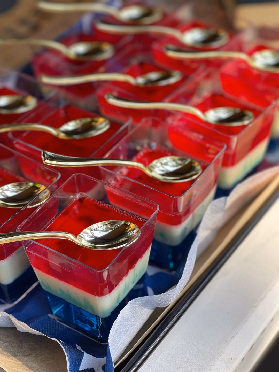 Rows of small clear cups holding red, white, and blue layered gelatin, each with a spoon resting on top, on a serving tray