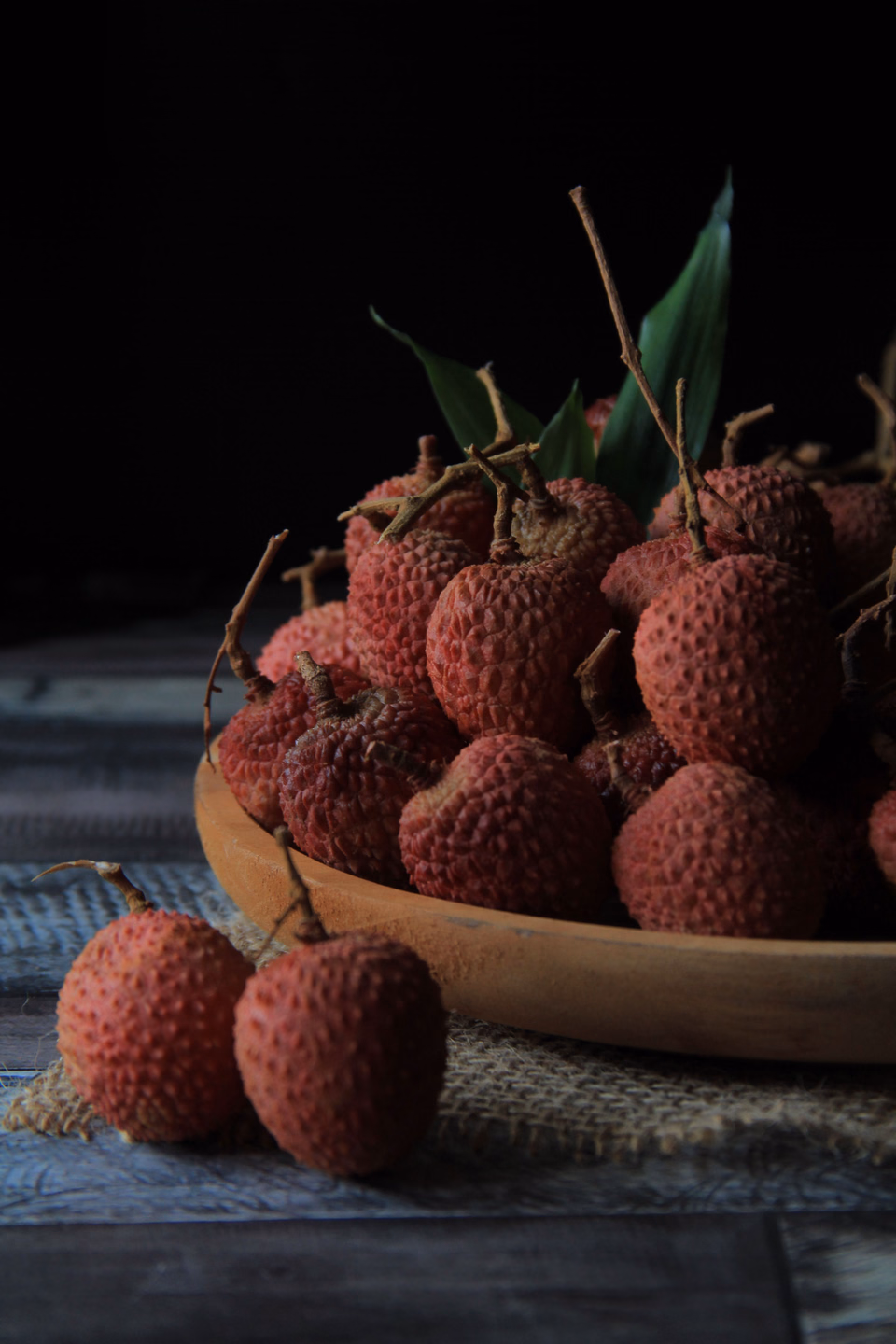 A wooden bowl piled with fresh red lychees in their bumpy shells, two more lying on the dark table in front