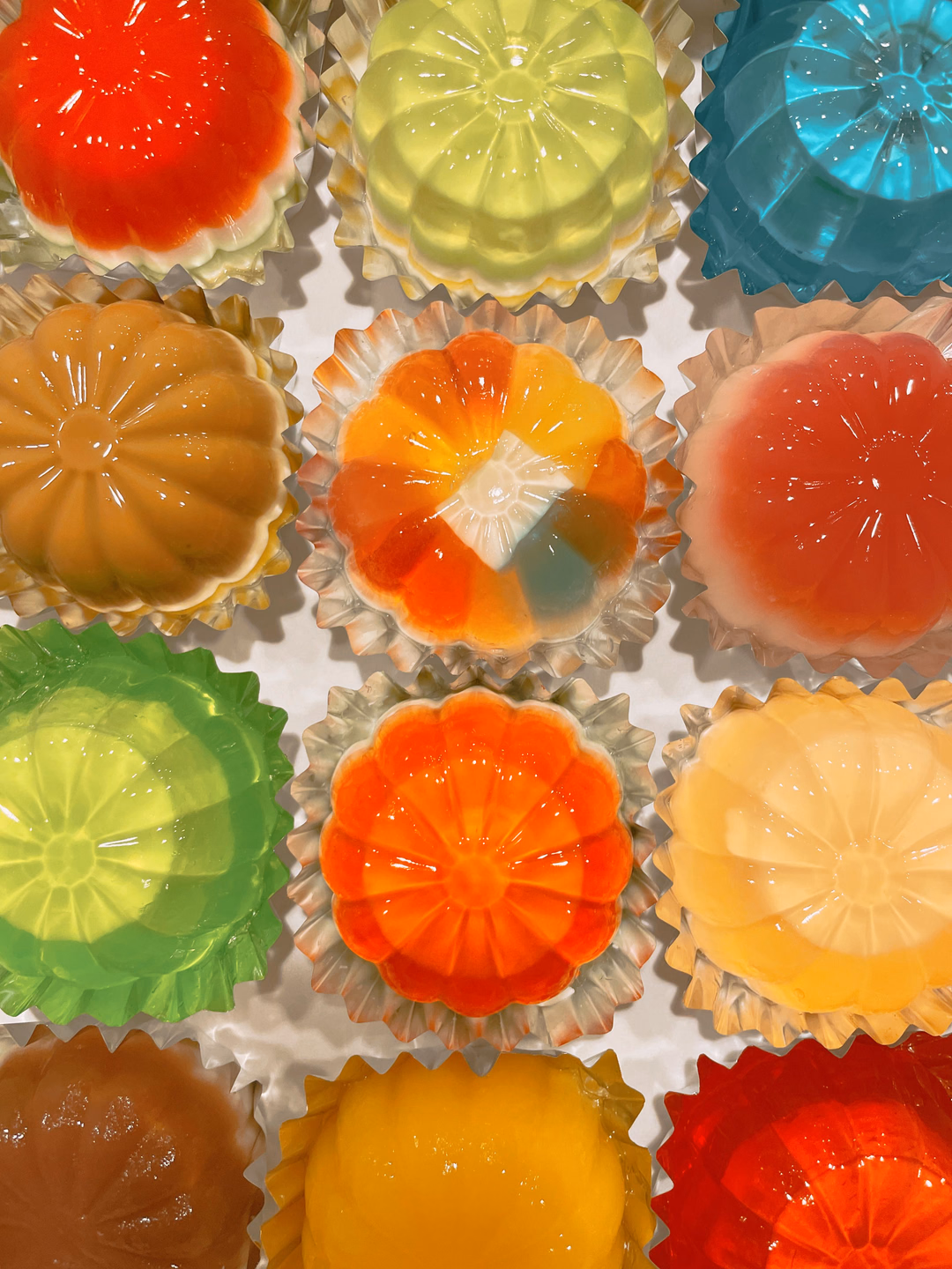 Top-down grid of molded gelatin domes in green, orange, red, yellow, and clear, each with a scalloped edge, sitting in paper cups