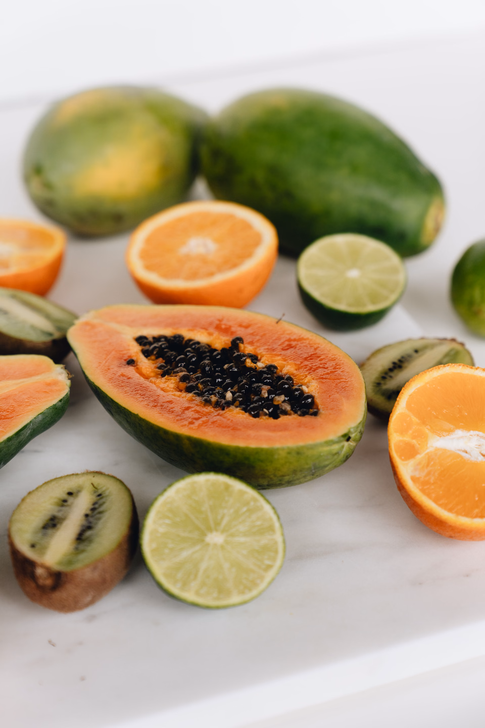 Halved papaya with its black seeds exposed, sliced kiwi, oranges, and limes on a white counter