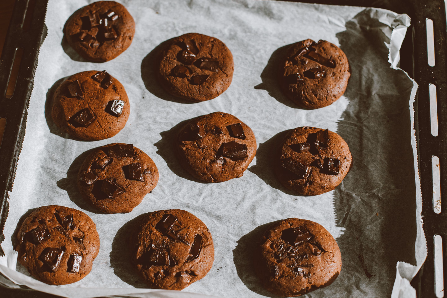 A dozen dark chocolate cookies studded with chocolate chunks on a parchment-lined baking sheet