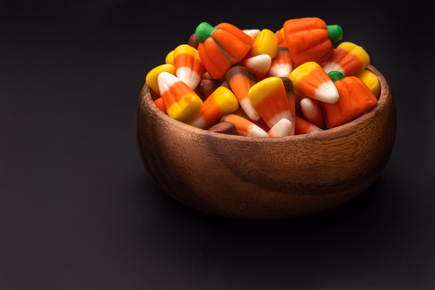 A small wooden bowl heaped with candy corn and candy pumpkins on a black background