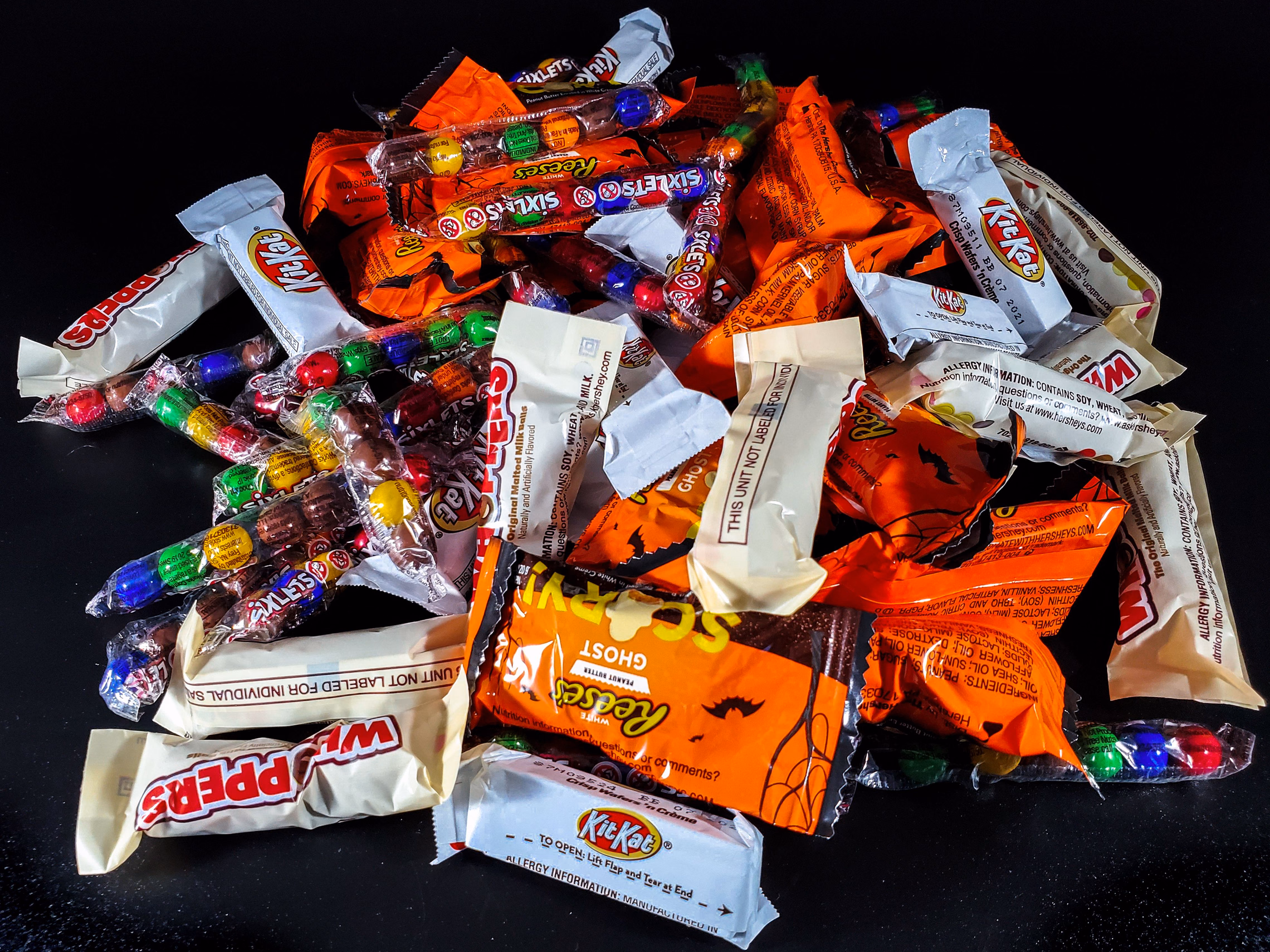 A pile of fun-size Halloween candy on a black table, including Kit Kats, Whoppers, Reese's ghosts and tubes of Sixlets