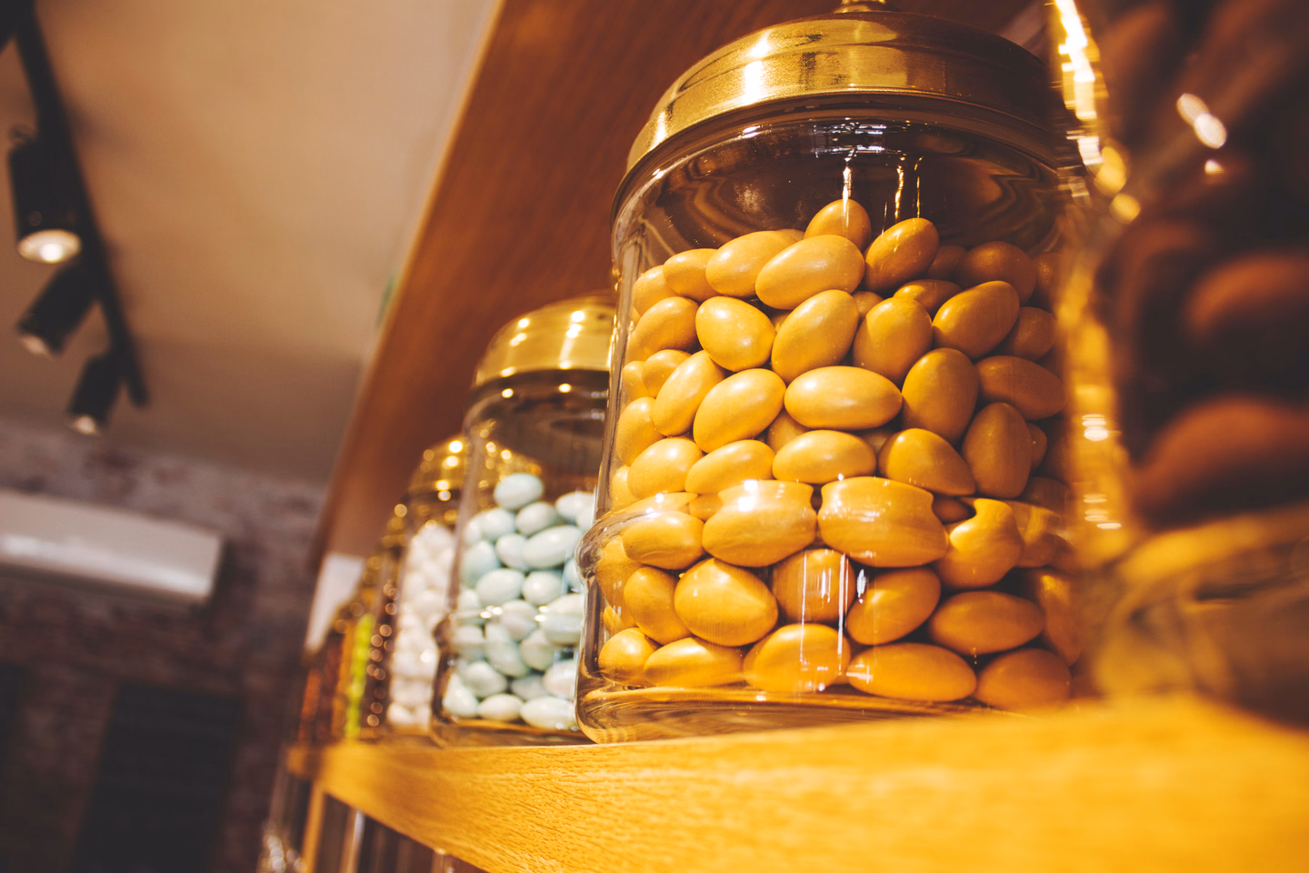 Tall glass jars with gold metal lids on a wooden shelf, filled with pale yellow and pale blue sugar-coated candies