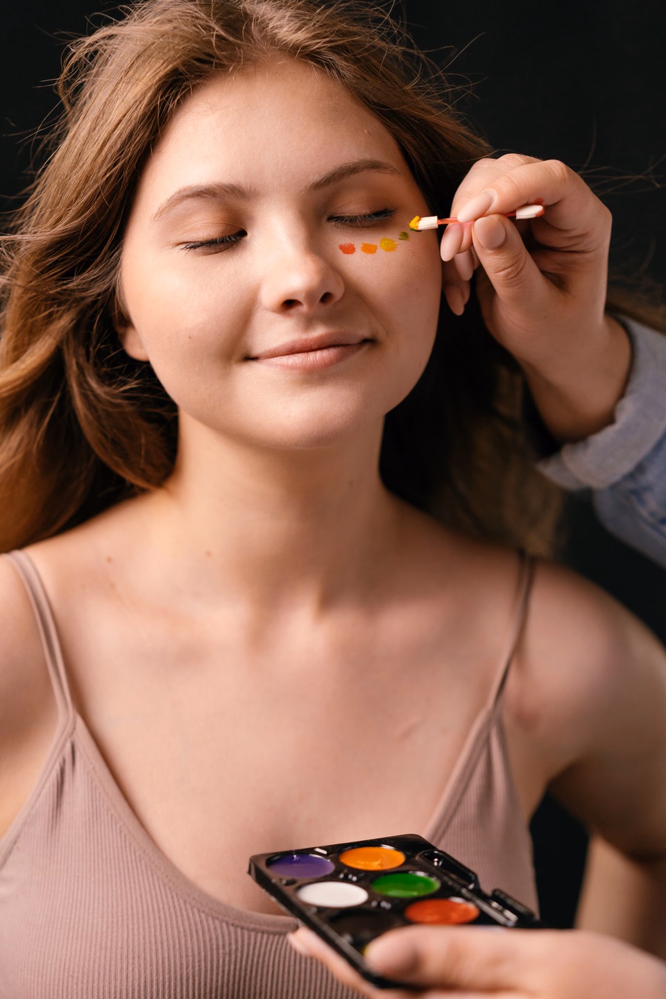Makeup artist dotting orange and yellow face paint onto a woman's cheekbone from a six-color cake palette while she keeps her eyes closed