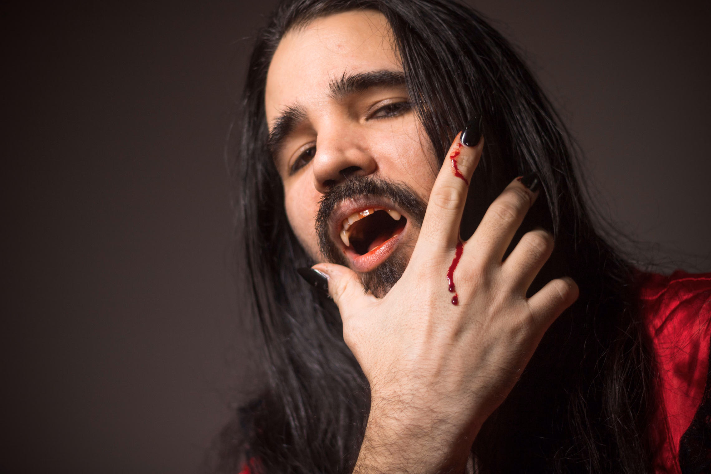 A man in vampire costume with fangs bared, fake blood at the corner of his mouth and running down two fingers of the hand raised to his face