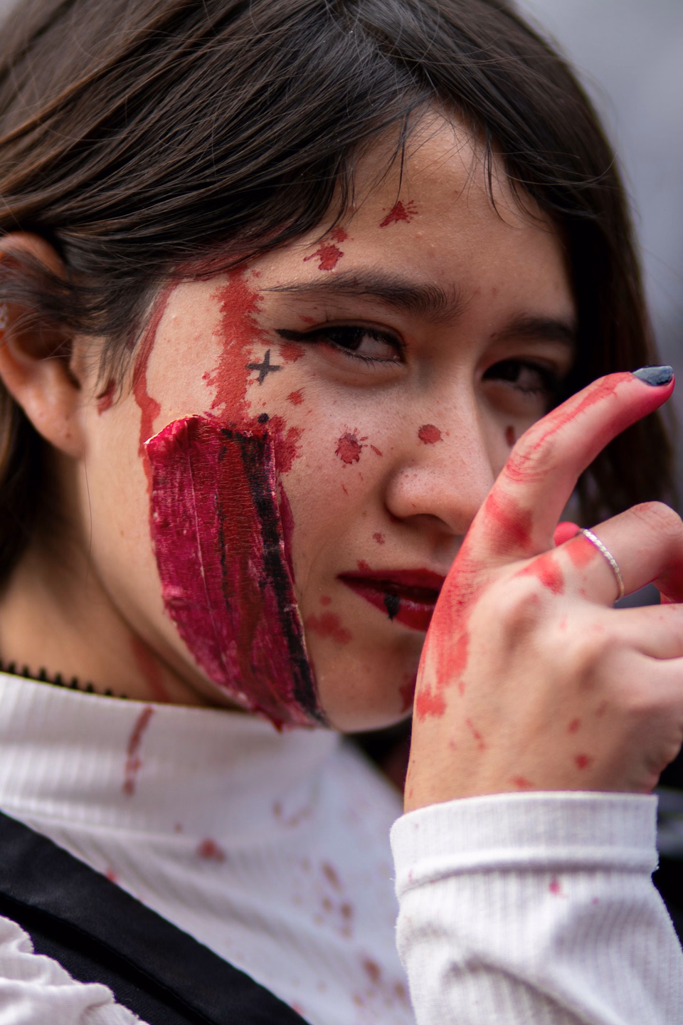 Young woman with a smeared stripe of dark red fake blood down one cheek, painted-on stitches, and blood on her fingers
