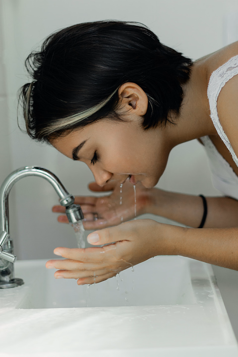 Woman bending over a white bathroom sink, rinsing her face with water from cupped hands