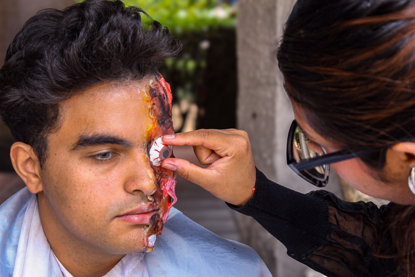 Makeup artist working on a raw, layered special effects wound that covers one side of a young man's face from temple to jaw