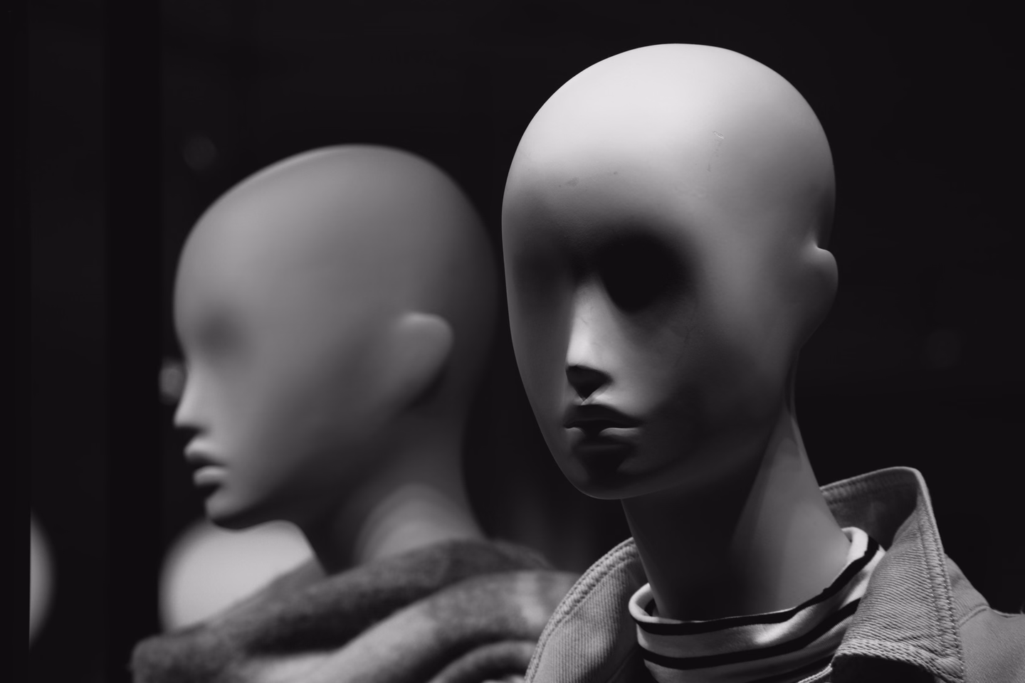 Black and white close-up of two bald mannequin heads with blank, softly sculpted faces, one wearing a denim jacket