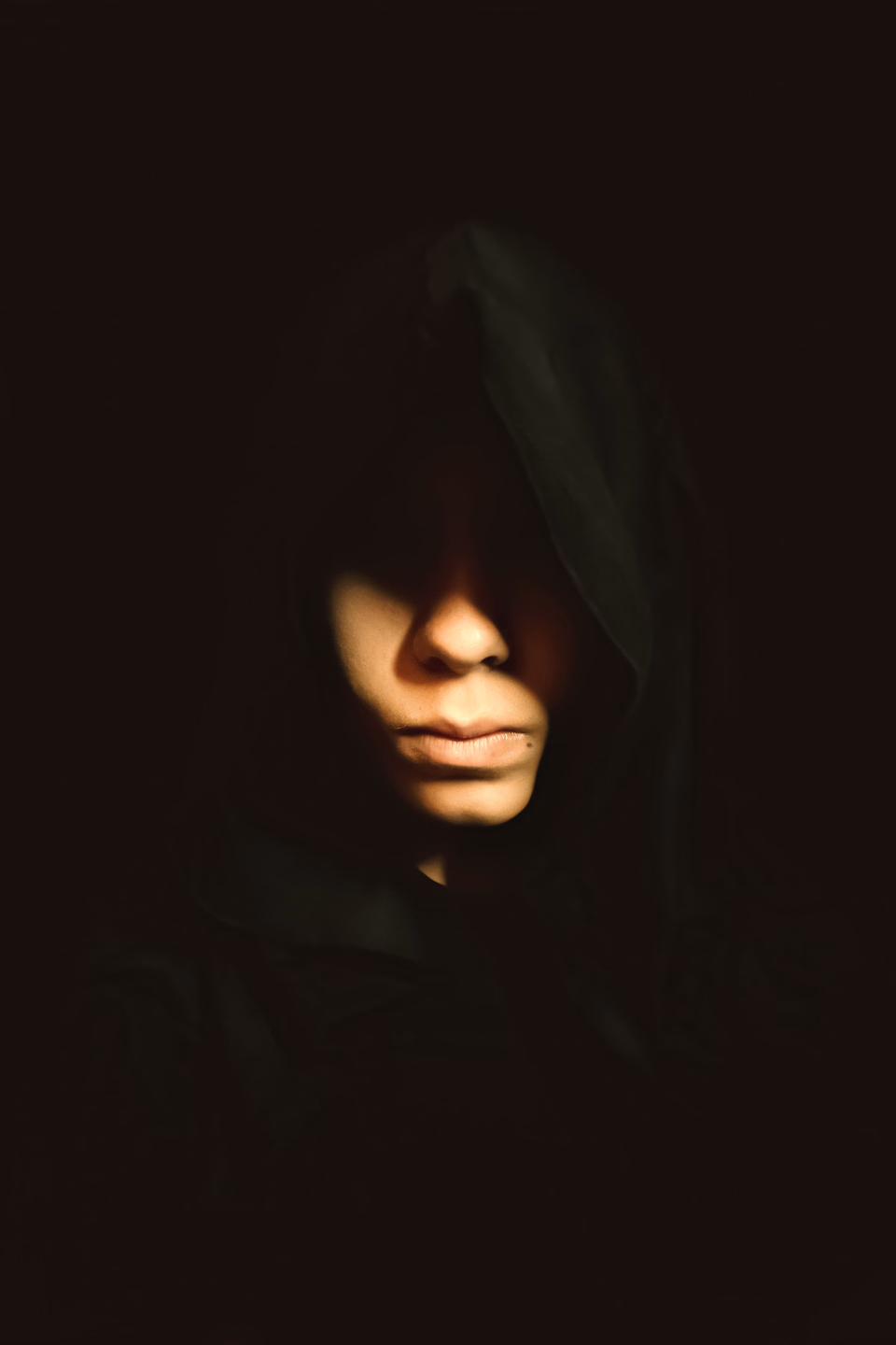 A figure in a dark hood against a black background, the eyes lost in shadow and only the nose, mouth and chin lit