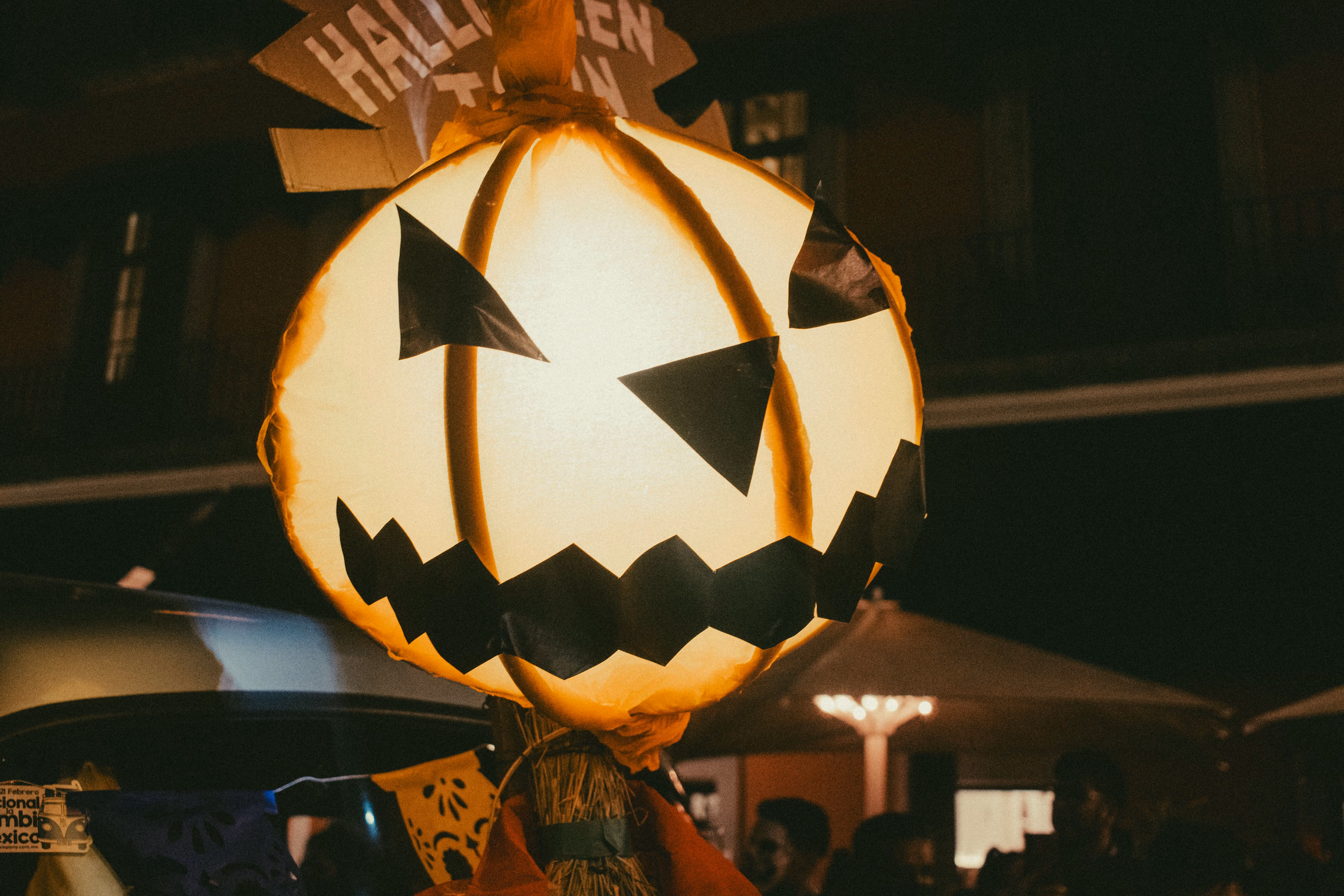 A round paper jack-o'-lantern glowing warm white from inside at night, with black paper triangle eyes and a zigzag mouth, a hand-painted Halloween sign above it and a crowd blurred below