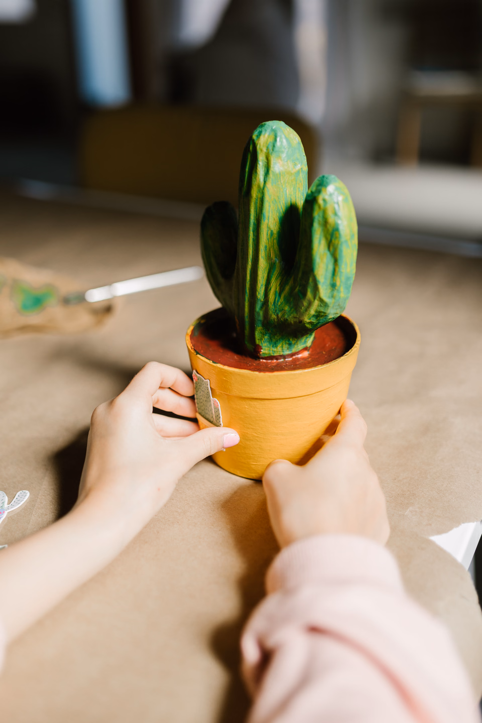 Two hands on a paper-covered table holding a small handmade green cactus sculpture with thick brush-painted ridges, planted in a yellow painted pot