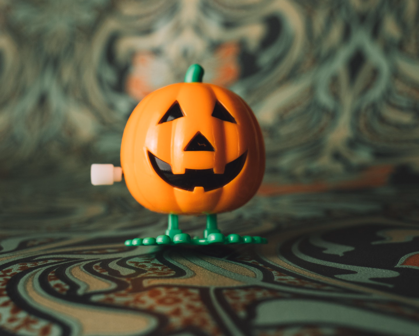 A glossy orange plastic wind-up jack-o'-lantern toy with black triangle eyes and a toothy grin, standing on green plastic feet on a swirling green patterned cloth