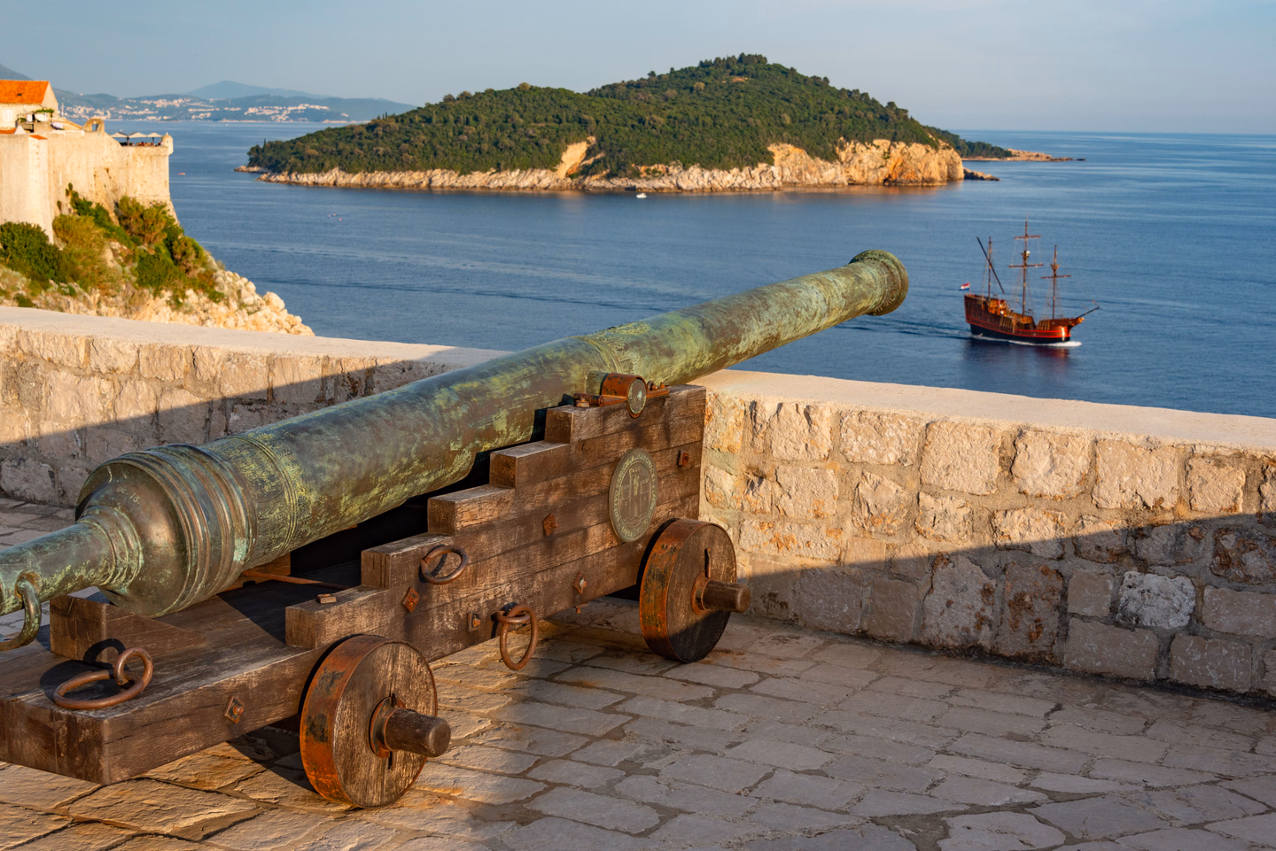 A green bronze cannon barrel on a wooden carriage with iron-rimmed wheels, aimed over a stone rampart toward the sea and a sailing ship