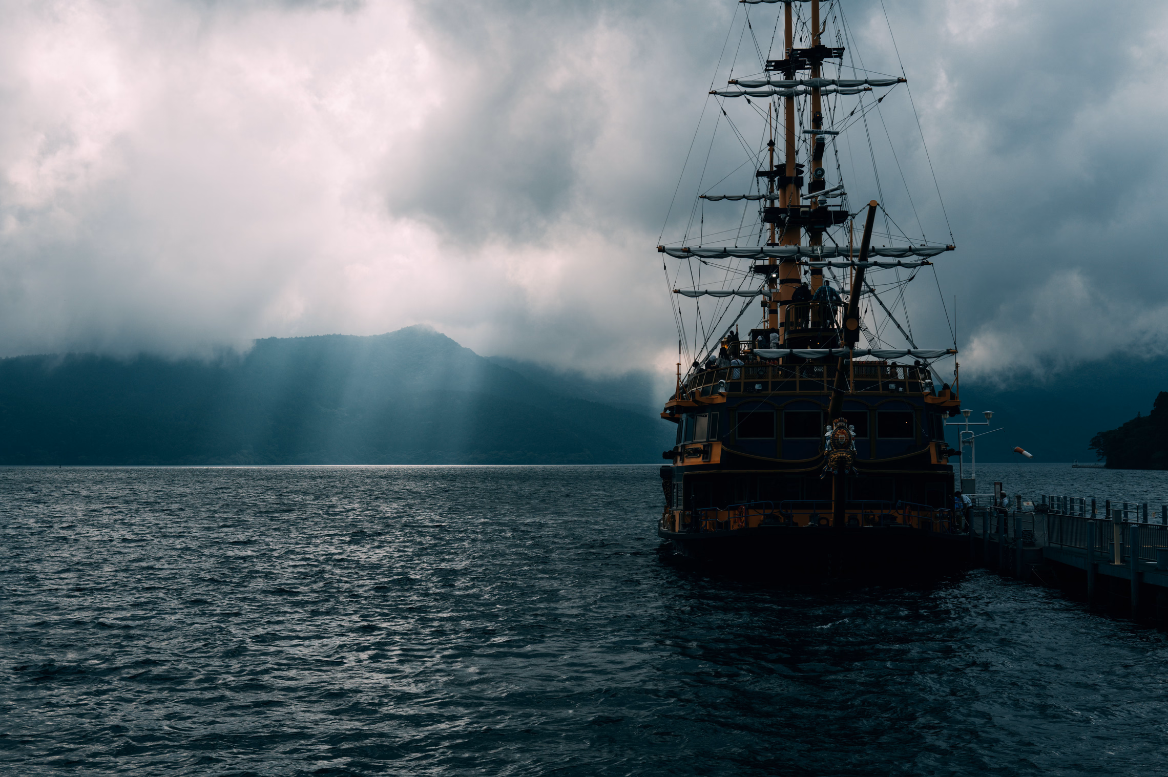 A tall-masted wooden sailing ship moored at a pier on dark gray water under heavy storm clouds, with a shaft of light breaking over the hills behind it