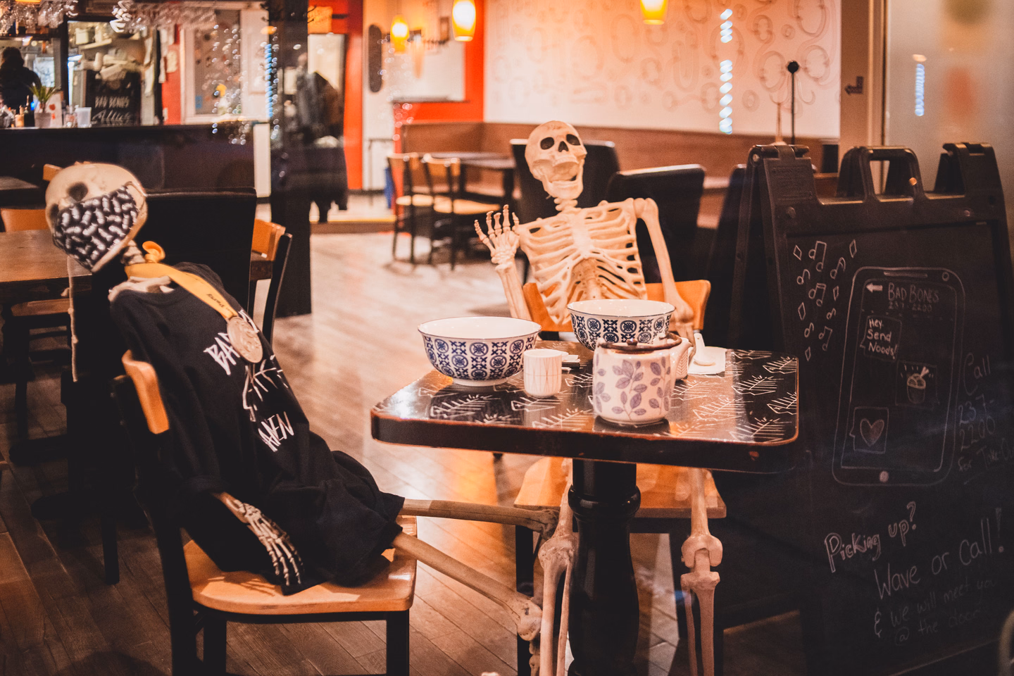 Two skeletons seated at a small cafe table set with patterned bowls and a teapot, one dressed in a black hoodie and slumped in its chair, the other upright with a hand raised