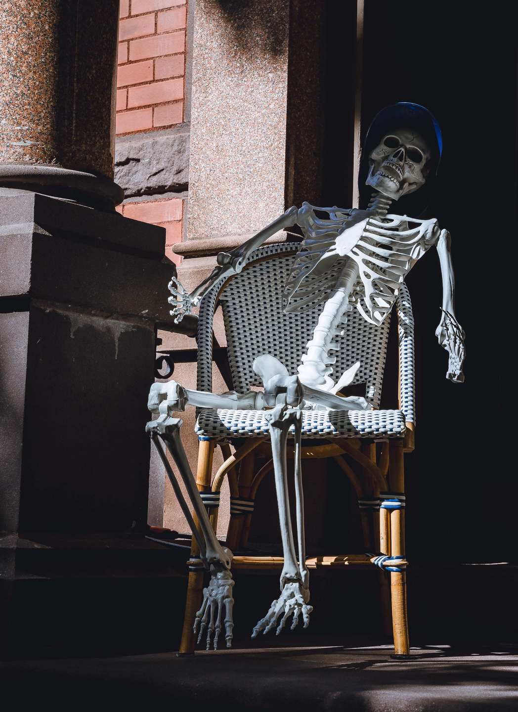 A skeleton in a blue cap sprawled in a woven bistro chair beside a stone column, one arm hooked over the chair back and legs stretched out
