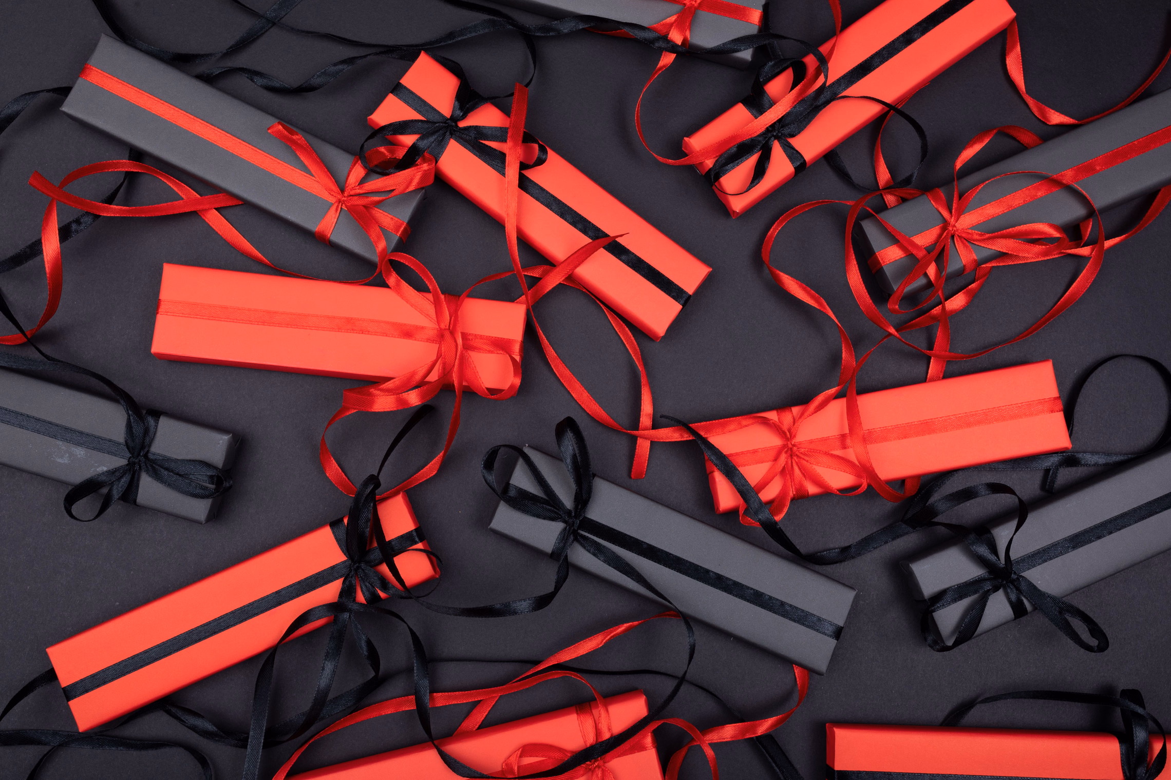 Long flat gift boxes wrapped in red and charcoal paper and tied with loose red and black satin ribbons, scattered across a black surface