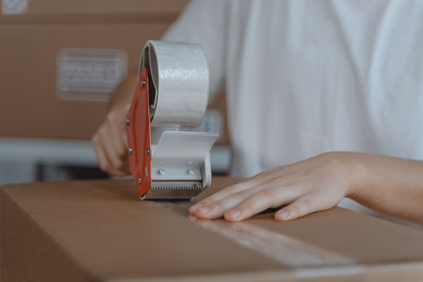 Close view of a hand holding a red tape gun, running clear packing tape along the seam of a plain cardboard box