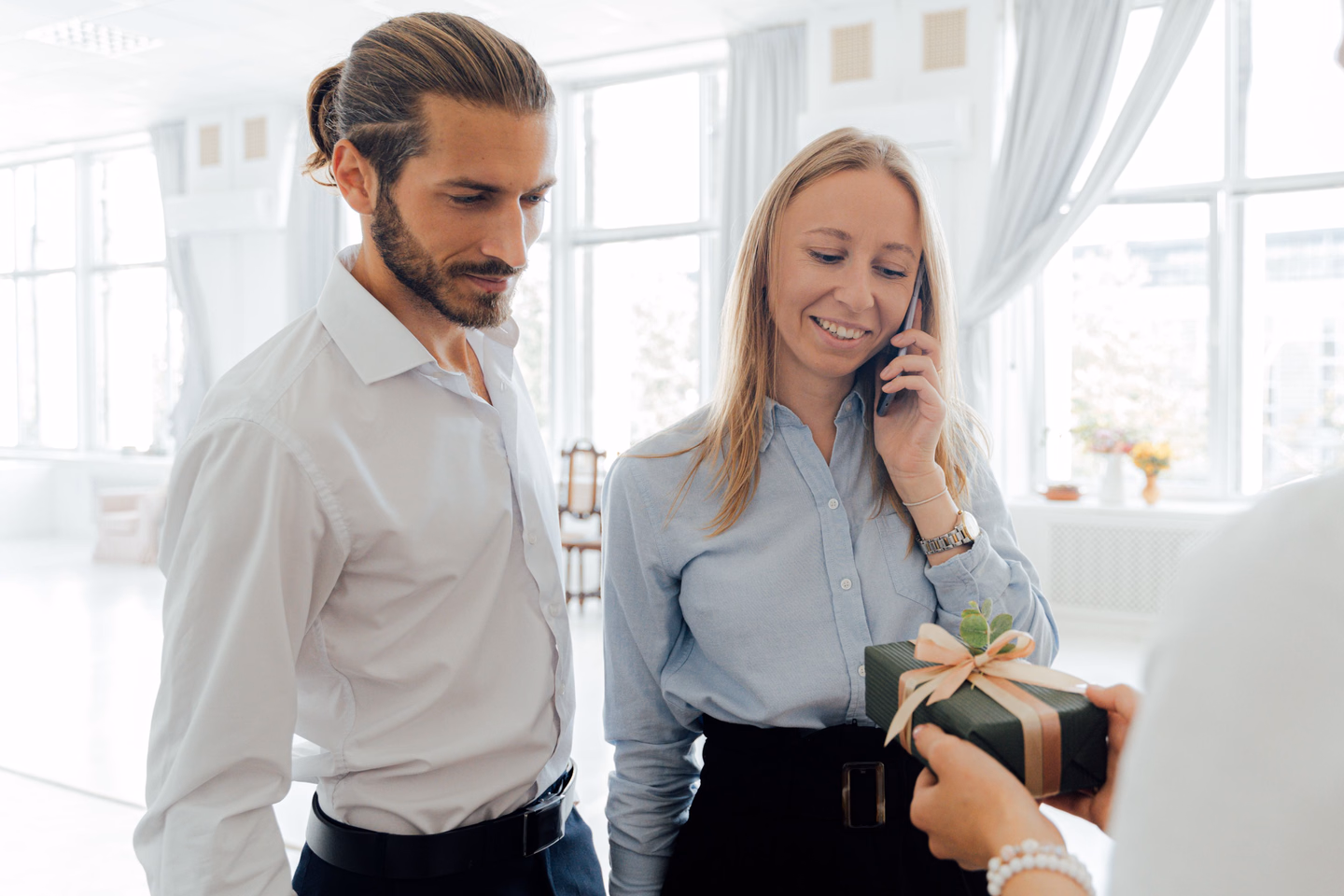 A smiling woman on a phone call in a bright white office is handed a small dark green gift box tied with a cream ribbon, while a bearded coworker in a white shirt looks on