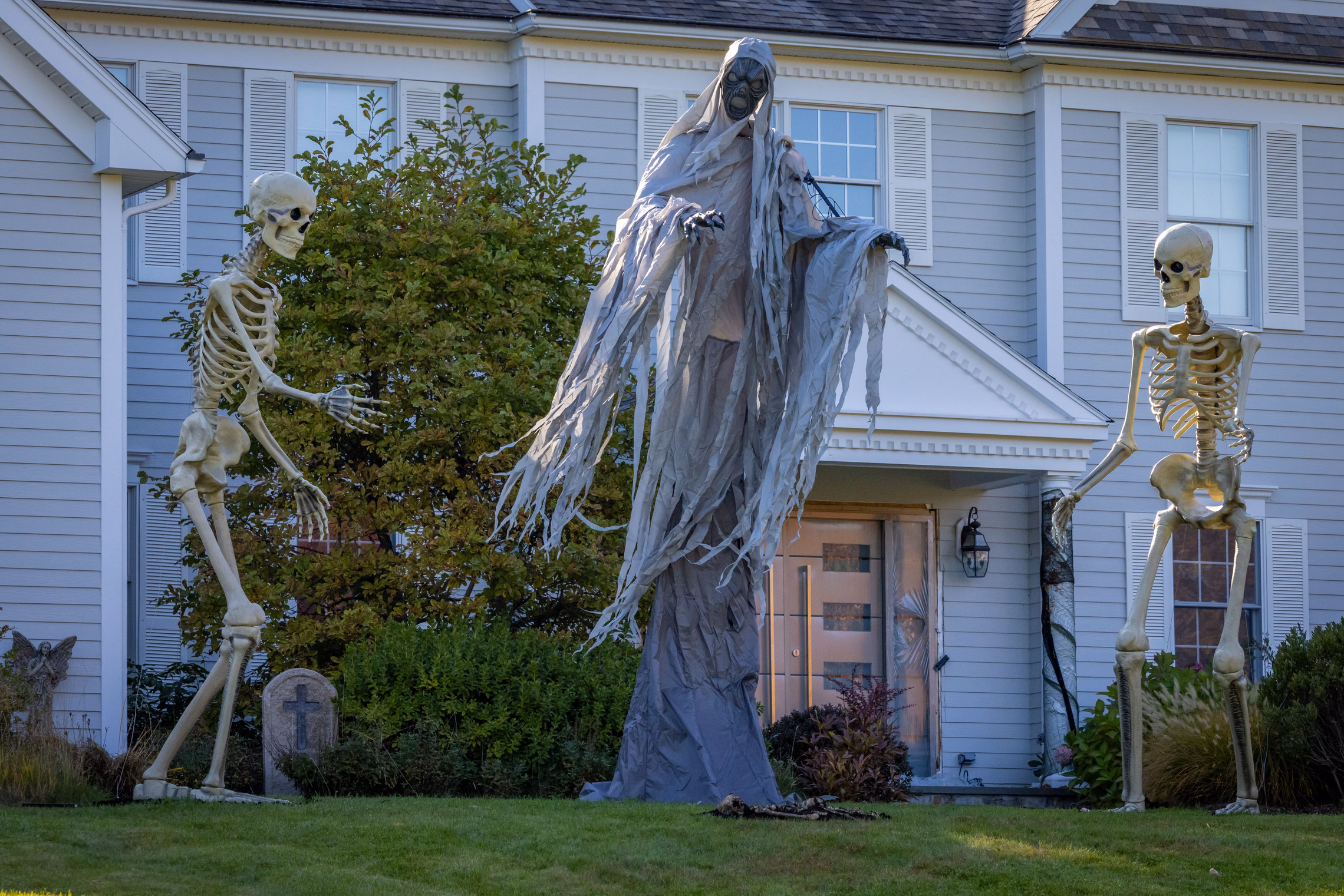 A front yard decorated for Halloween with two tall skeletons, a hooded reaper figure and a tombstone on the lawn