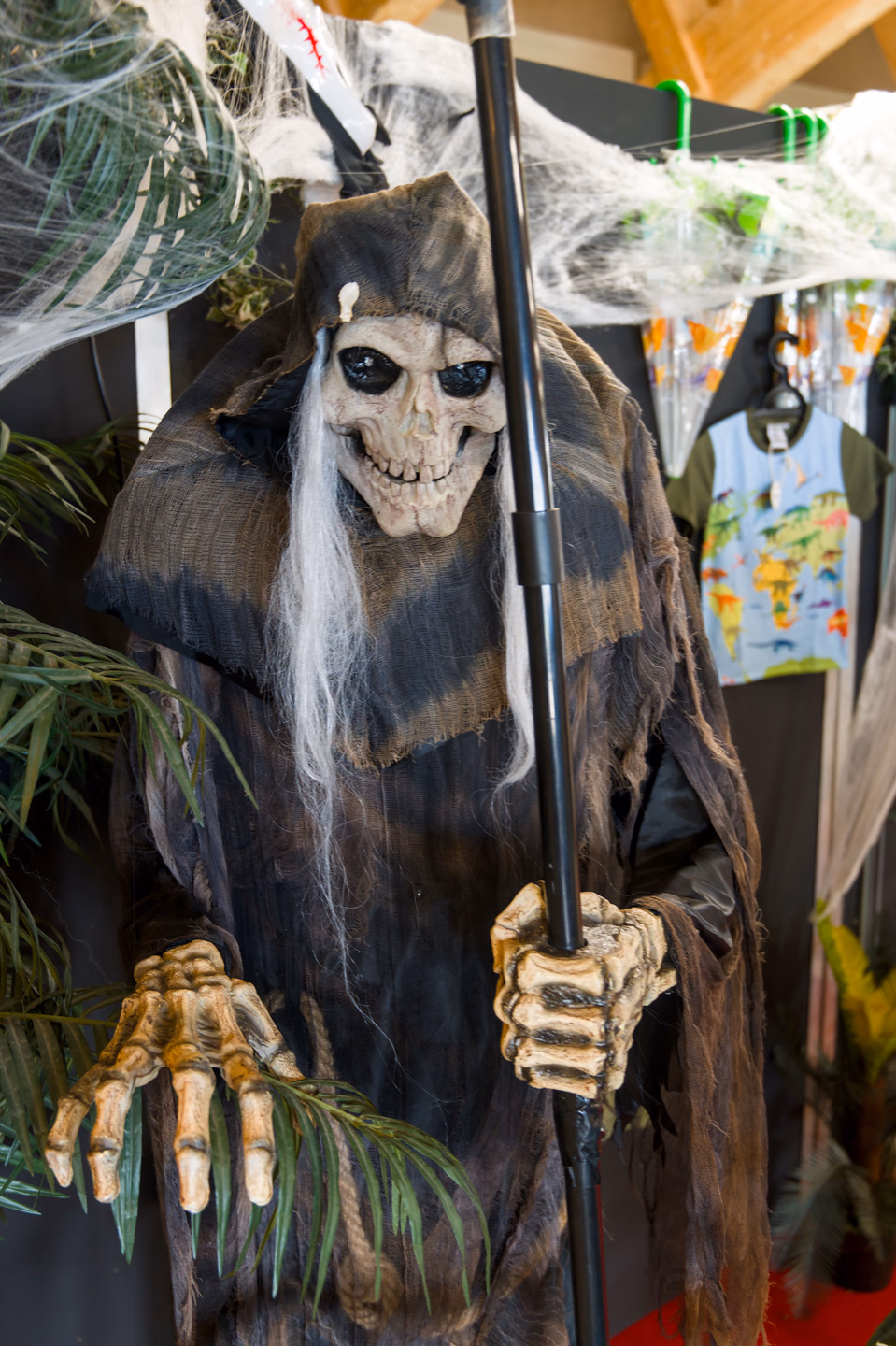 A hooded reaper decoration with a skull face, long white hair and bony hands gripping a scythe, draped in fake cobwebs