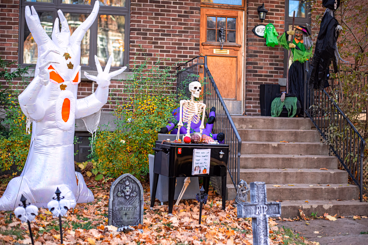 A brick house's front steps decorated with an inflatable ghost, a seated skeleton, a witch figure and tombstones in fallen leaves