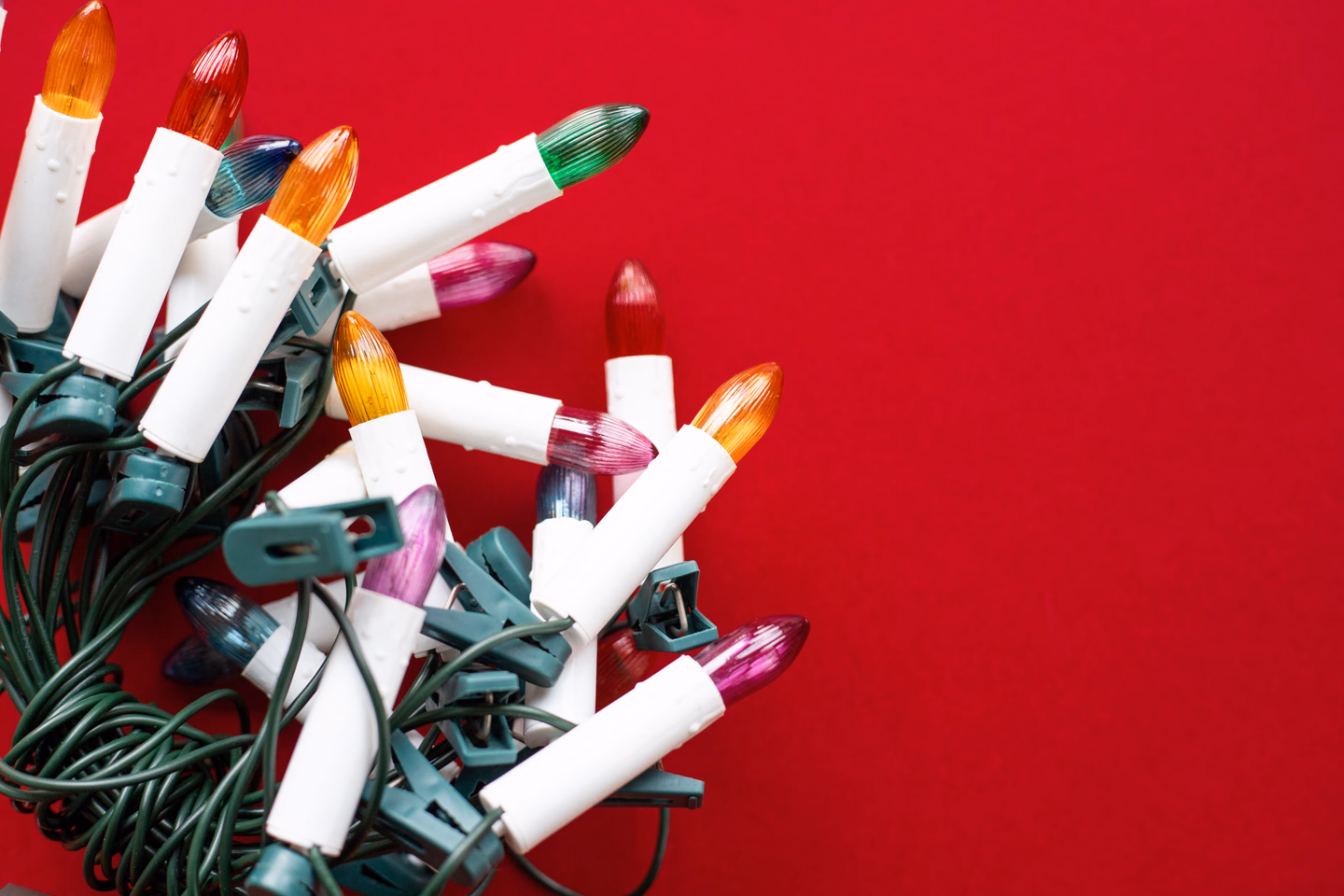 A tangle of old-style candle bulb lights with white plastic sockets and colored glass bulbs on green wire, against a red background