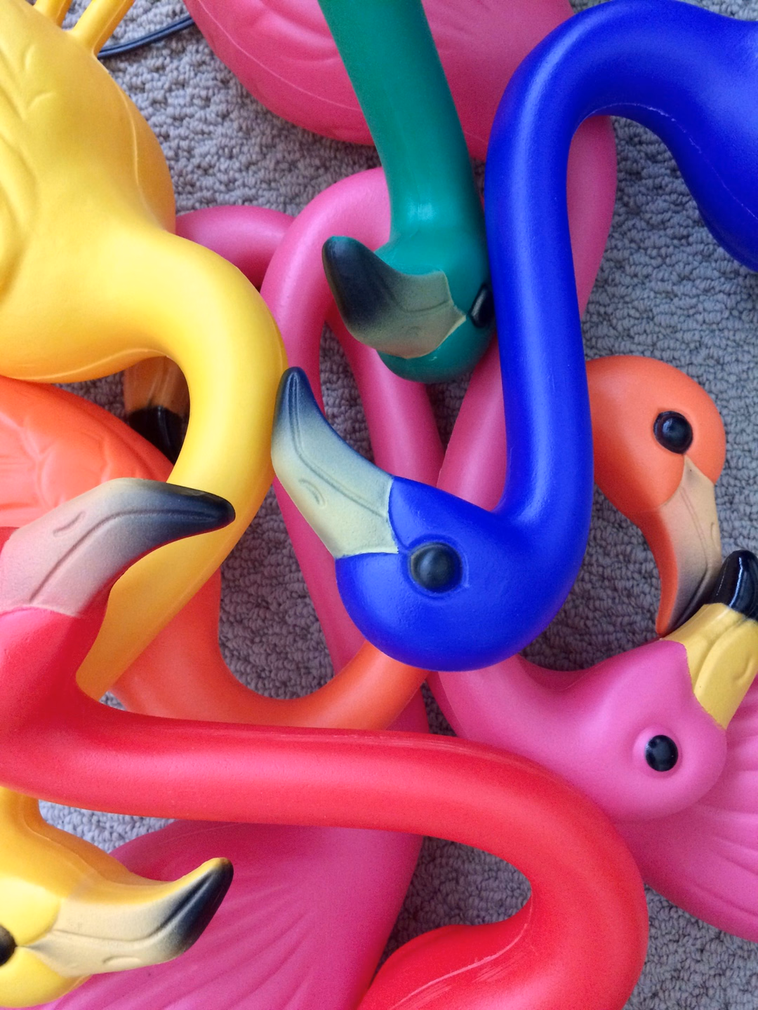 A jumble of hollow plastic flamingo lawn ornaments in pink, yellow, cobalt blue, green and coral, with molded eyes and black-tipped beaks