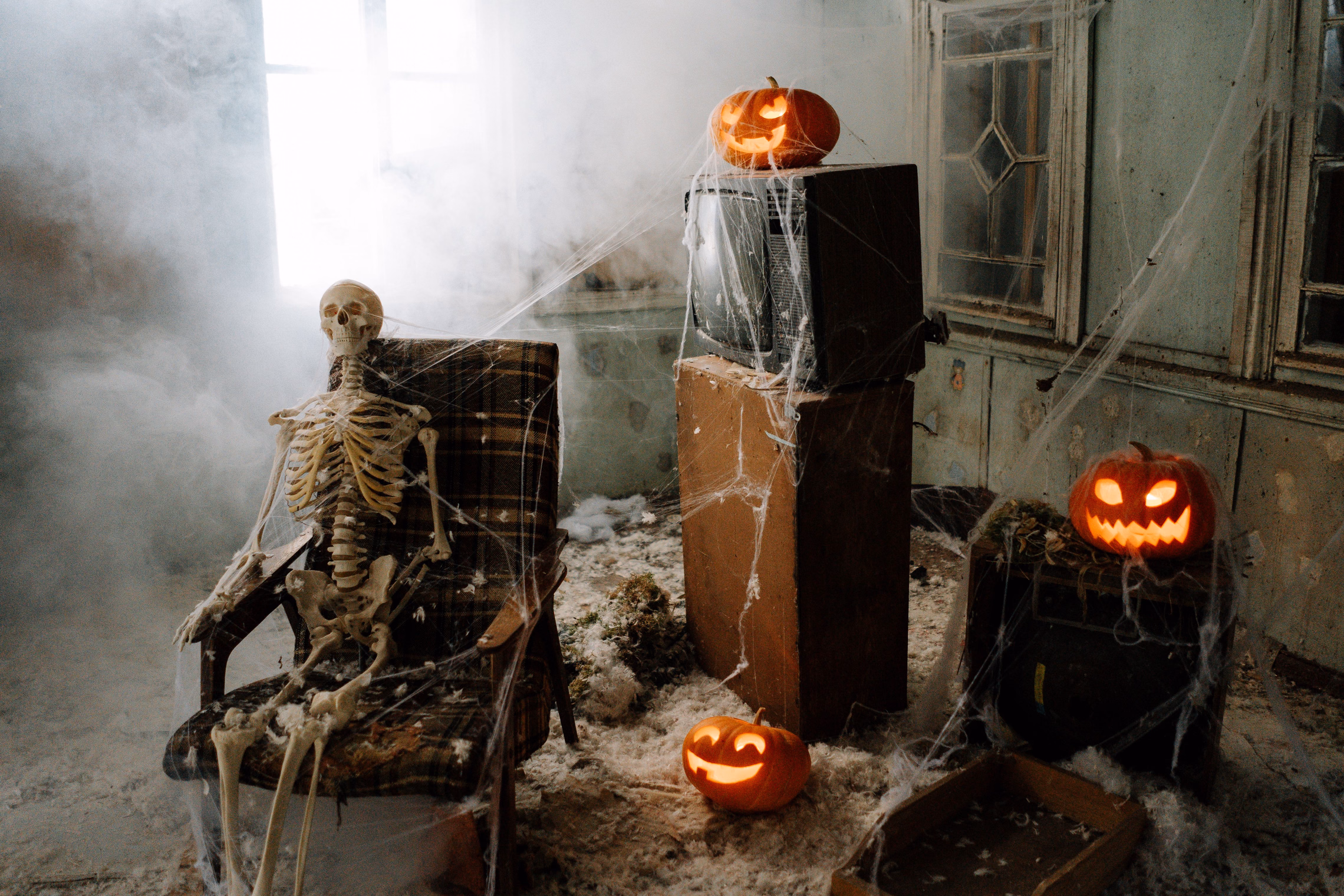 A skeleton slumped in a plaid armchair in a fog-filled room, with jack-o'-lanterns stacked on an old television set on a wooden cabinet