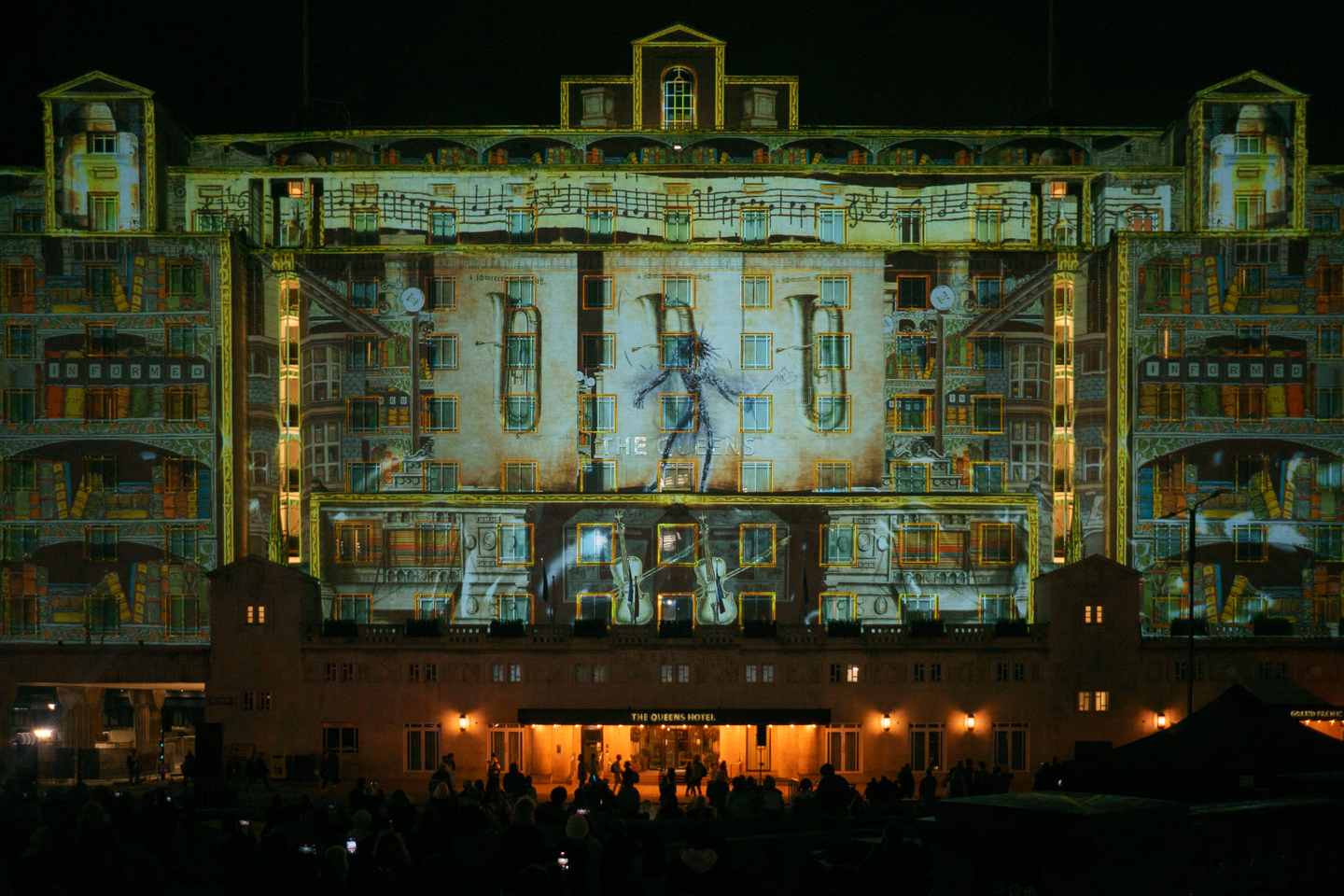 A large hotel facade at night covered edge to edge in projected artwork, with a crowd in silhouette in the foreground