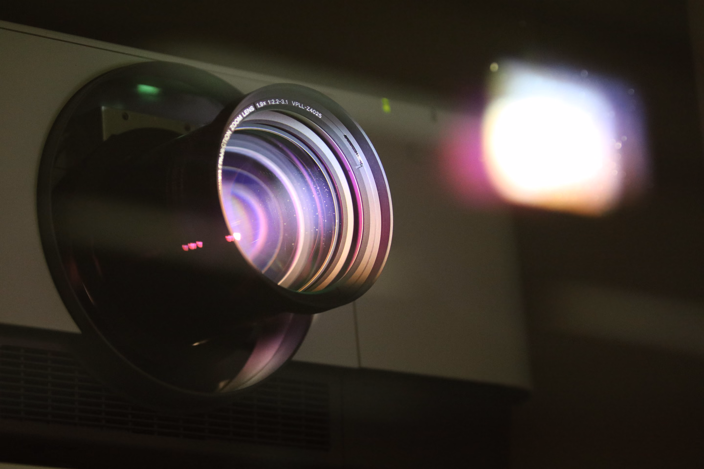 Close view of a projector's zoom lens in a dark room, with the bright square of its light source glowing out of focus behind it
