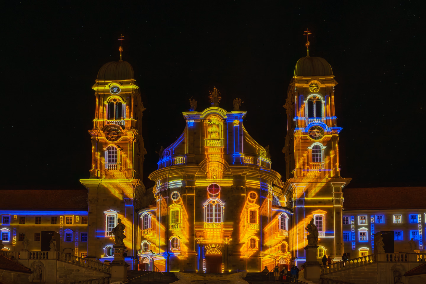 A baroque abbey with two towers at night, its windows, columns and cornices picked out in projected gold and blue light