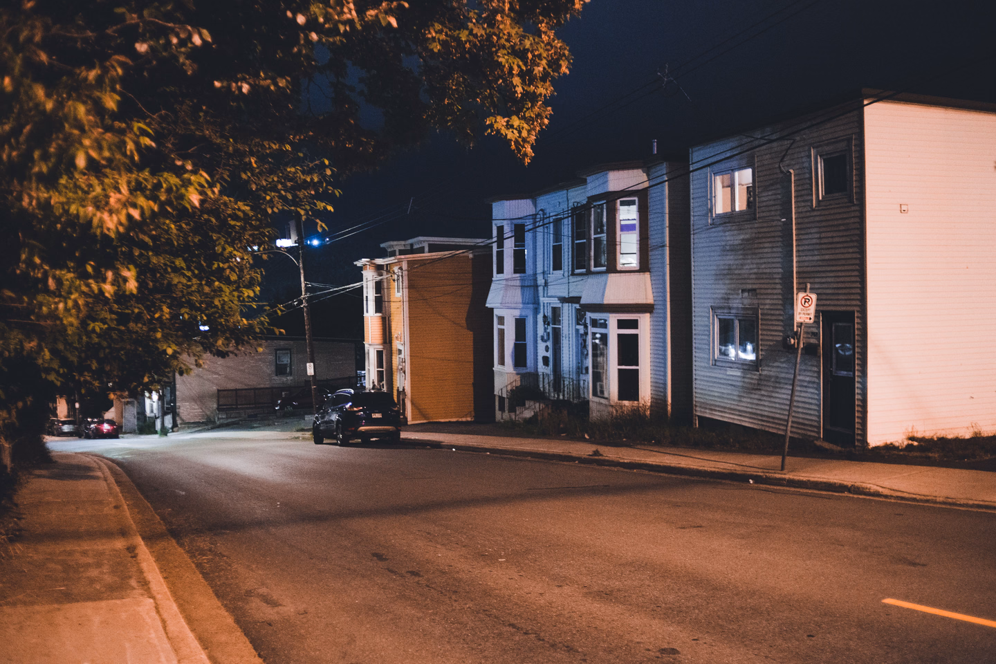 A residential street at night with a row of clapboard houses, a parked car and a single streetlight