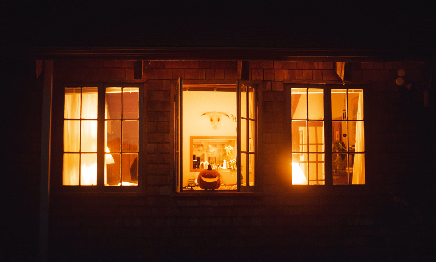 Three lit windows in a shingled house at night, with a carved jack-o'-lantern sitting in the open middle window