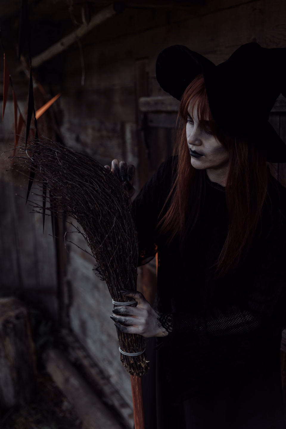 A witch in a black pointed hat and dark cloak stands in a shadowy wooden shed, gripping a bundled twig broom