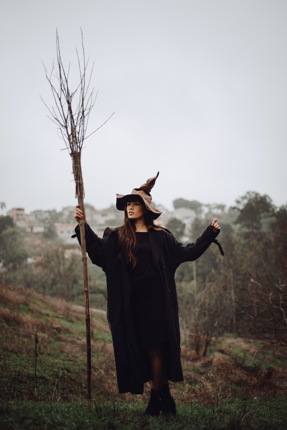 A witch in a long black cloak and pointed hat holds a tall twig broom on a grassy hill under a gray sky