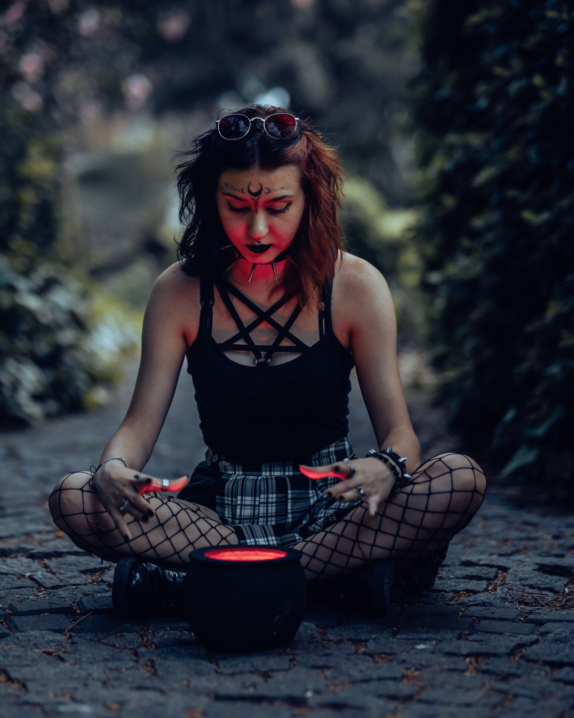 A witch in red face paint sits cross-legged on a cobblestone path, hands raised over a small cauldron glowing red