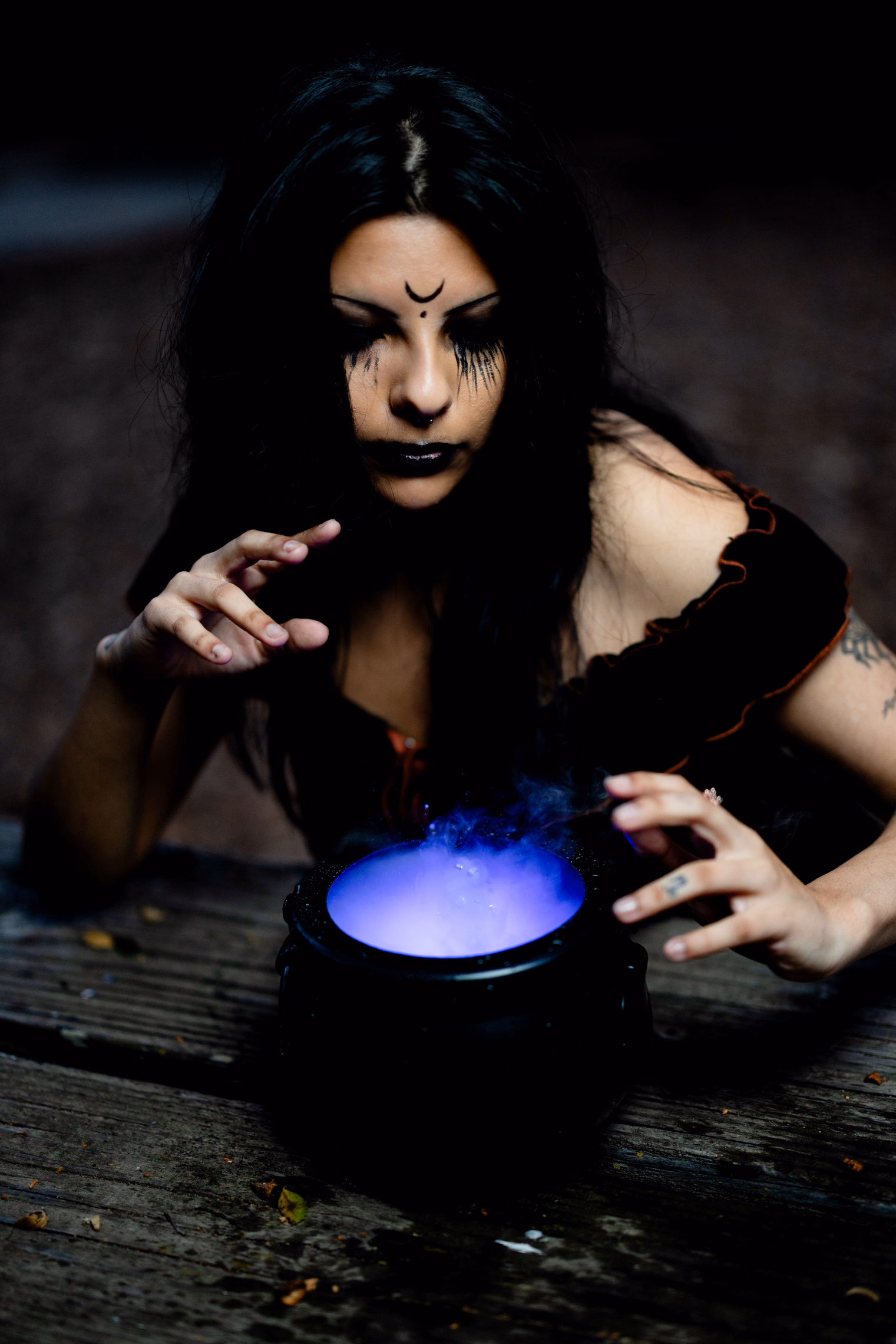 A dark-haired witch with a painted crescent on her forehead leans over a small black cauldron glowing violet from inside