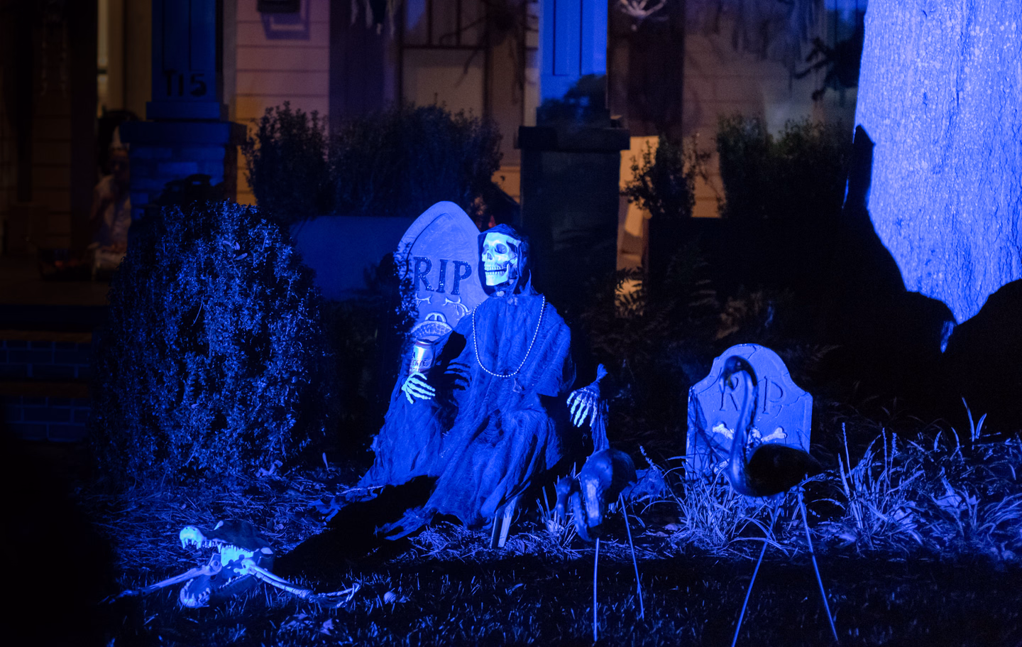 A seated skeleton reaper and two RIP tombstones on a front lawn at night, lit deep blue from the ground