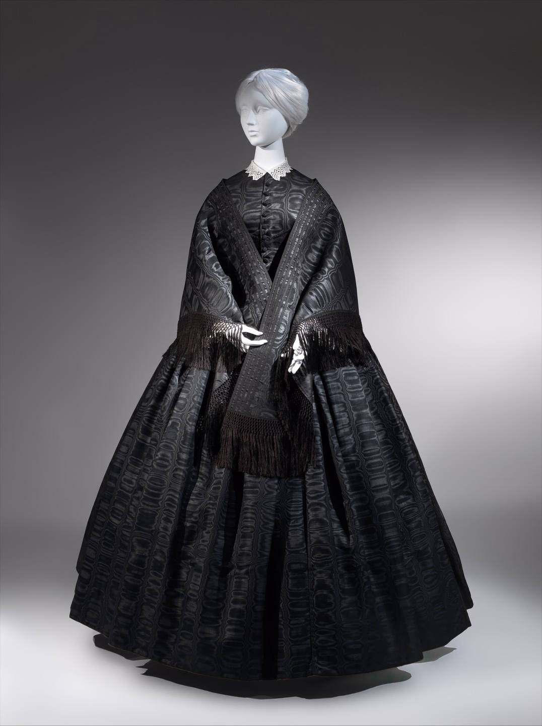 A black mourning ensemble on a mannequin: a full crinoline skirt and fitted bodice in black silk with a wavy watered pattern, a small white lace collar, and a long black shawl with deep fringe draped over the shoulders and hanging to the knees