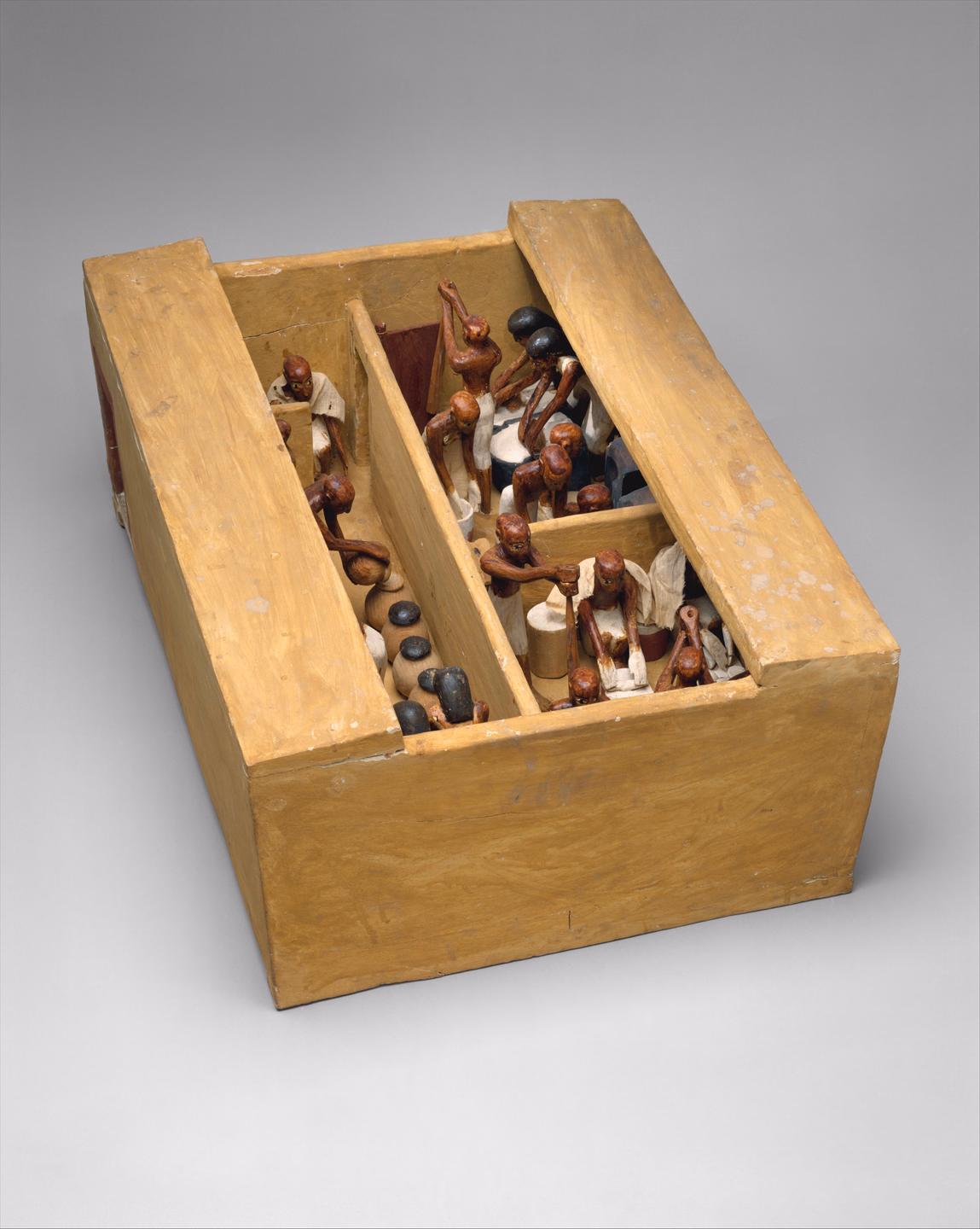 A painted wooden model of a small open-roofed building divided into two rooms, crowded with small red-brown figures at work grinding, kneading and tending tall dark jars