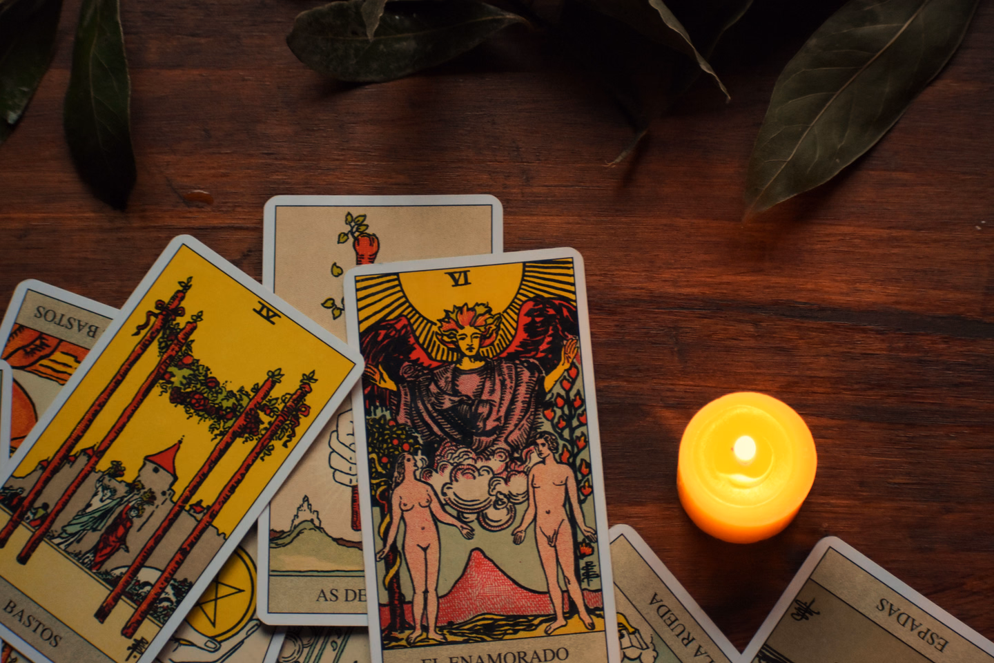 Rider-Waite style tarot cards, a Spanish-language edition showing The Lovers, fanned across a dark wooden table beside a lit candle and a few bay leaves