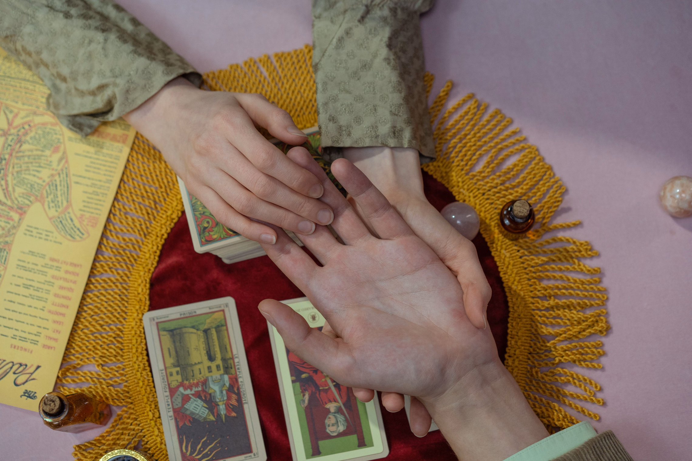 Overhead view of a palm reading: a reader's fingers trace the lines of an open palm over a red cloth with gold fringe, tarot cards and a printed palmistry chart beside them